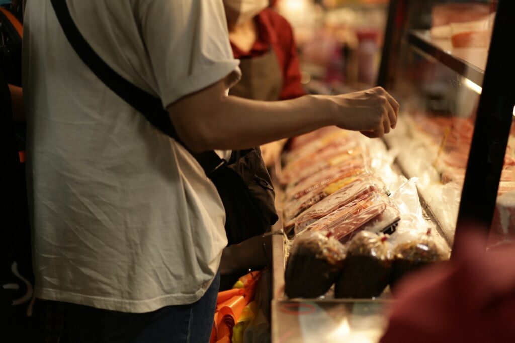 Person in a white shirt browses a deli counter filled with packaged meats. A shop attendant in the background. Warm lighting creates a cozy feel.