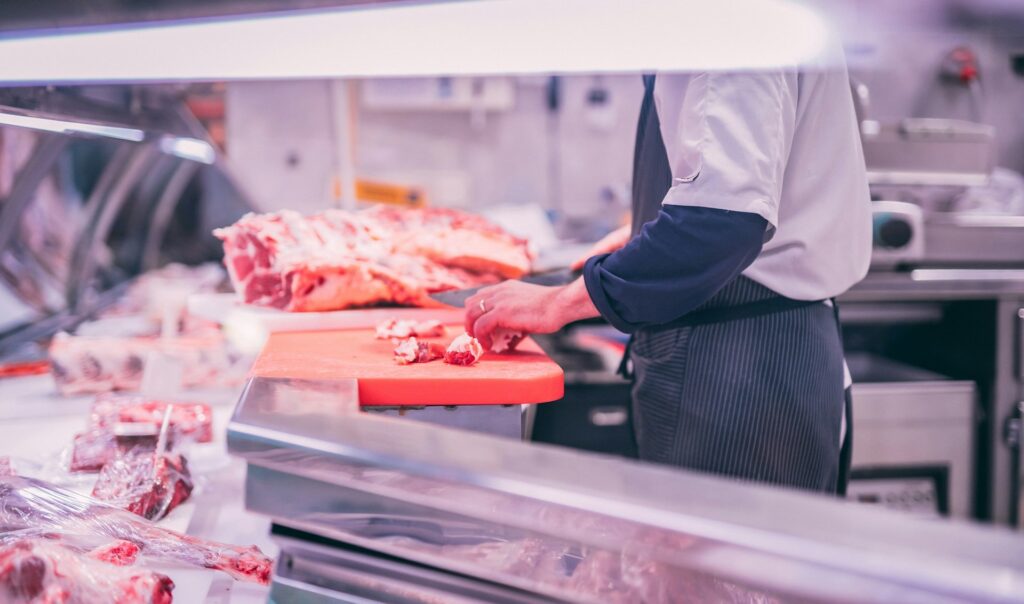 A butcher cuts raw meat on a red board at a meat counter. The setting is clean, with large cuts of meat displayed. The atmosphere is professional.