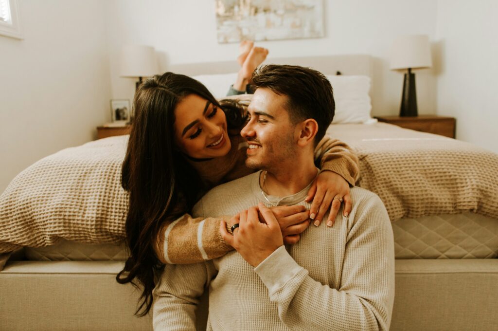 A smiling woman lying on a bed on her stomach with her arms around a smiling man sitting against the foot of the bed
